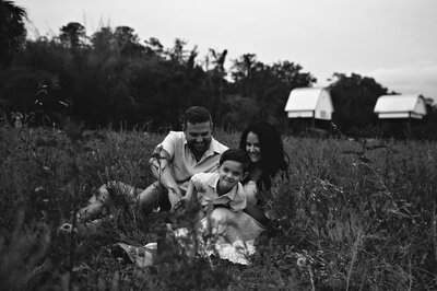 Black and white photo of a family sitting in a wildflower field with bat houses in the background, captured during a joyful outdoor session in Gainesville Florida by Salt + Heart Photography