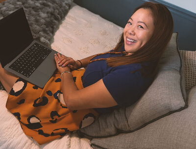 An Asian-American woman sitting on a bed with a laptop on her lap, turning and smiling.