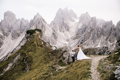 Groom is assisting the Bride down a dirt hiking trail with an impressive, spiky mountain range rising behind them.