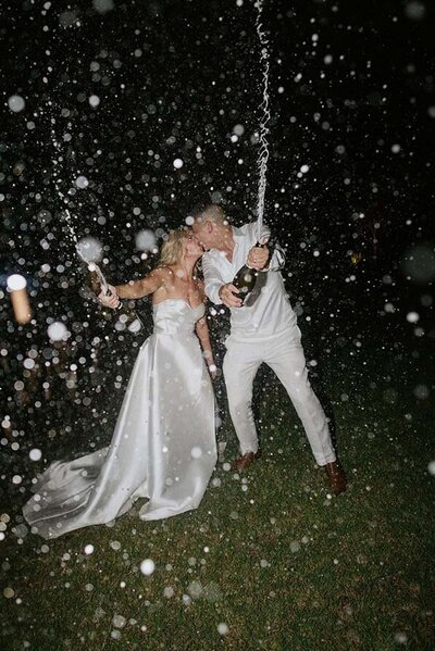 Bride and groom in white outfits spraying champagne and kissing under a flurry of bubbles during an outdoor Melbourne celebration at night.