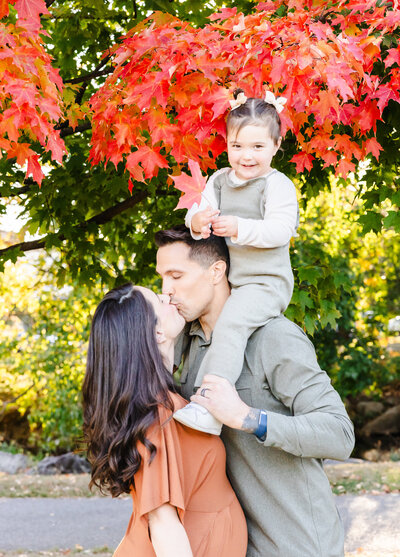 Lifestyle photo of laughing daughter on father's shoulders while mom and dad kiss at an Essex County, New Jersey park in the fall.