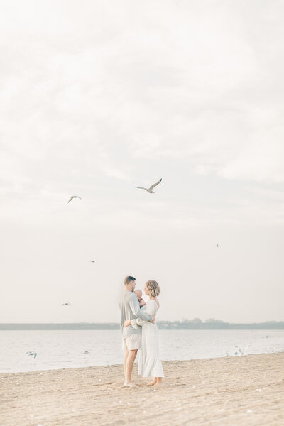 Family picture of a mother and father hugging their 6-month-old-son on the shore of a Kansas City beach while seagulls fly overhead