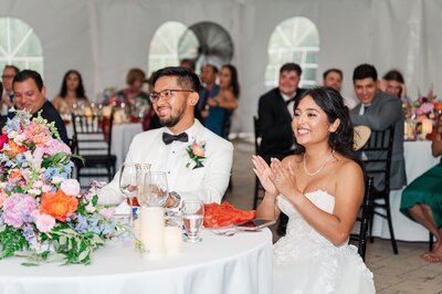 A bride and groom smile and clap at their wedding reception. 