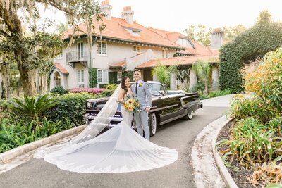 Bride and groom standing in front of a vintage car surrounded by green plants and brick home