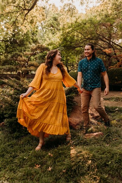 Girl in yellow flowy dress leads guy in button up shirt down garden path surrounded by greenry in Fort Worth Botanical Garden
