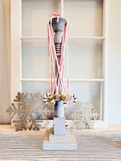Vintage-inspired glitter bell ornament from Sweet Pea Ink Creative, featuring blush pink and gold ribbon accents, jingle bells, and candy-striped details. Displayed on a spindle-style stand with glitter snowflake decor in the background.