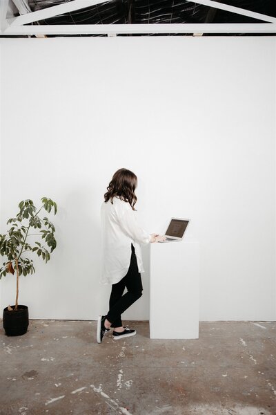A professional woman in a minimalist studio, standing confidently at a laptop atop a white podium, surrounded by a touch of greenery, symbolizing focus, growth, and the seamless virtual assistance services offered by KP Virtual Assistance for photographers