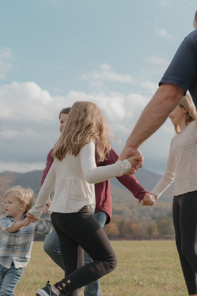 A family of five play ring around the rosey together in a field with a mountain in the background. The sky is blue with white clouds in it. 