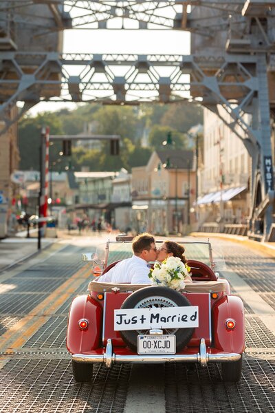 Newlyweds kiss as they drive over the Mystic Bridge in their vintage car.