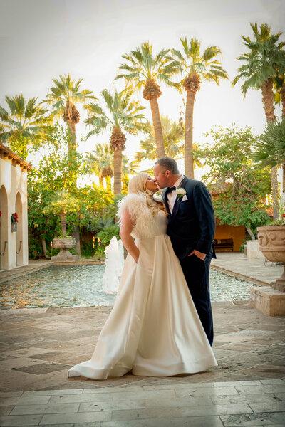 Bride and groom kiss in front of pool surrounded by palm trees