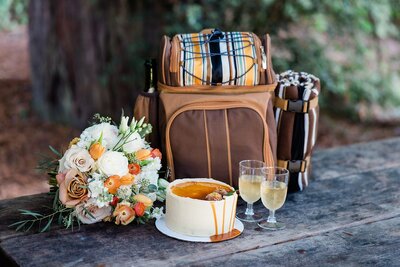 A small wedding cake, backpack and wedding bouquet sits on a picnic table.