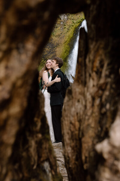 Bride and groom embracing in front of a waterfall as seen through a hole in a tree.