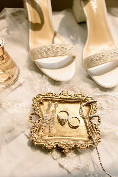 Flatlay of wedding rings, vintage tray, perfume, and bridal heels on tulle during a Charleston wedding day.