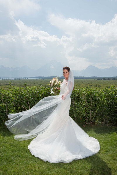 Bride at Jackson Lake Lodge Wedding – A bride in an elegant white wedding gown holds a bouquet as her flowing veil catches the wind on the lush lawn of Jackson Lake Lodge. The Grand Tetons create a stunning wedding backdrop under a bright sky.