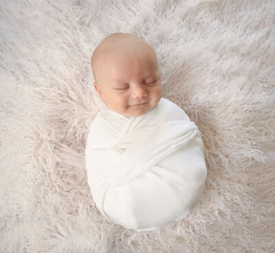 E. Benecki Photography photographed this in-home newborn session with the baby wrapped in white and snuggled in fluffy white furs while smiling.