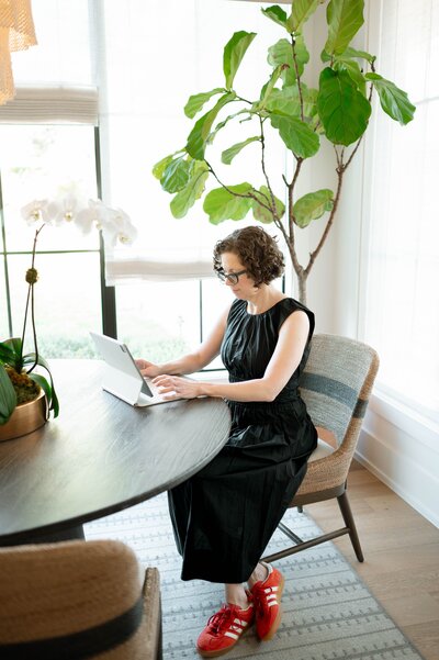 The owner of Scout & Bimble focuses on her tablet at a dining table, surrounded by soft natural light from floor-to-ceiling windows.