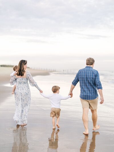 A family of four wearing classic blue colors walking on the beach holding hands.