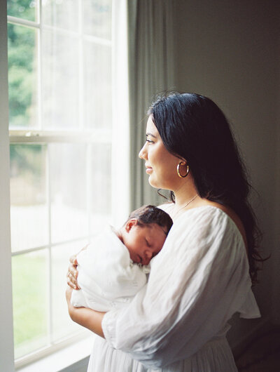 A mother holding her newborn while looking out of a window at a Richmond newborn photographer session by Katie Stansfield Photography.