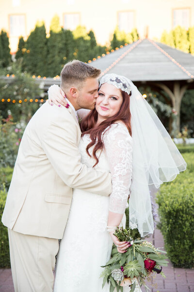 groom in a cream suit kissing his redheaded bride in a lush green garden