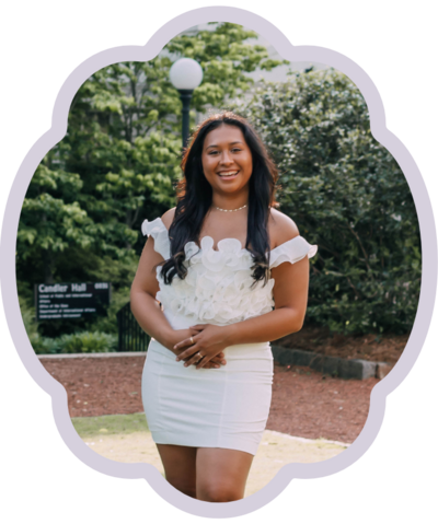 dark haired girl with tan wearing white ruffly dress smiling at camera in front of green trees