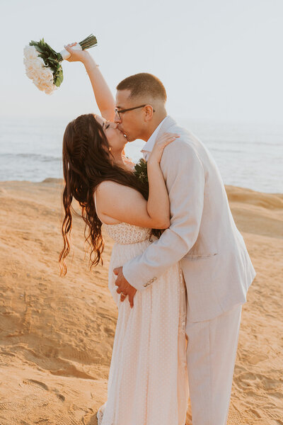 Bride and groom kissing and holding up a flower bouquet on Sunset Cliffs  in San Diego.