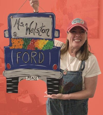 Woman in overalls and white shirt holding up wooden door hanger shaped like truck against red background