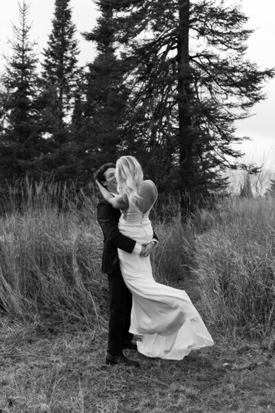 Eloping couple shares a playful moment as groom lifts bride among tall pine trees.