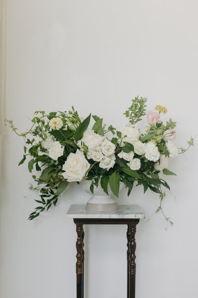 Elegant white floral arrangement displayed on a pedestal, featuring lush garden roses, spray roses, zinnias, and soft greenery in a modern compote vase, styled against a clean white wall—ideal inspiration for timeless wedding centerpieces and luxury floral design.