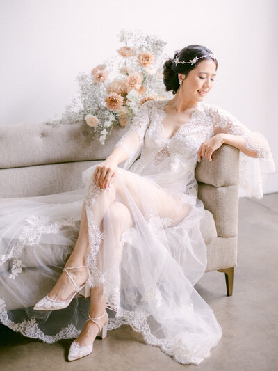 Beautiful bride in lace wedding dress leaning on sofa surrounded by floral decor and wearing floral headpiece