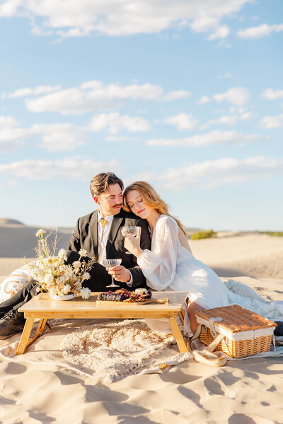 Bride and groom celebrating their elopement with a romantic picnic in the stunning sand dunes of Utah, surrounded by golden sands and a beautiful sky.
