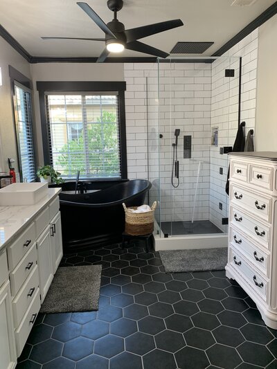 A large black soaking tub in a remodeled bathroom with hexagon black tiles, a white vanity, and subway tiled shower.