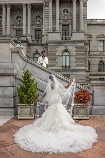 Black bride standing in front of a black groom standing on a stairwell wearing a white tuxedo in front of a historic grey building in Washington dc on wedding day