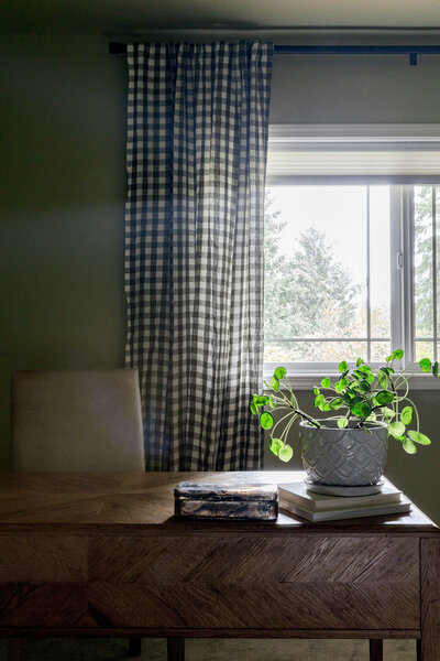 A wooden desk with books and a houseplant sitting on top of it, and open windows behind it with checkered curtains.