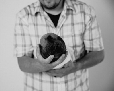 A newborn baby wrapped snugly in a white blanket, gazing wide-eyed at the camera, set against a minimalist white background.