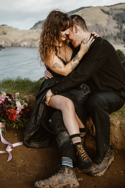 A couple in all black wedding attire snuggles up on a coastal cliff-side during their Oregon coast elopement.