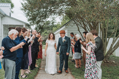 A bride in a lace wedding dress and a groom in a grey vest and turquoise tie share a kiss outdoors at the Michaux-St. James Foundation. The bride's veil flows gracefully around them, and the background features lush green trees. Shot by Quinn Photography LLC.