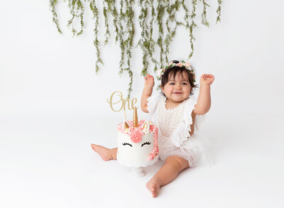 Baby girl in a white lace romper sits with a unicorn cake between her legs for her first birthday cake smash photoshoot. She is smiling and has her hands raise in the air with excitement.