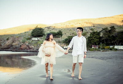 A pregnant couple stands at the beach, holding hands and looking at each other.