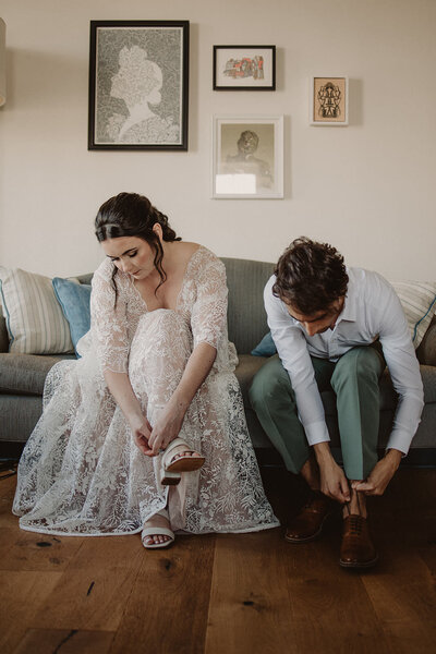 A wedding couple sitting on a couch together as they put on shoes 