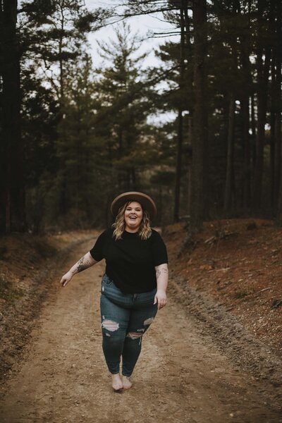 A fat woman out in nature, surrounded by trees, laughs and walks toward the camera. She is wearing a round felt hat, black t-shirt, and distressed skinny jeans.  