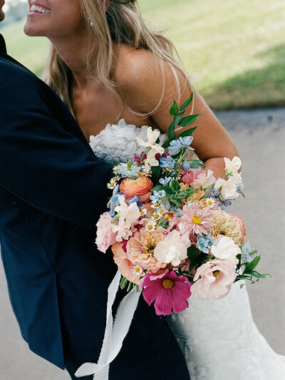 Bride in white lace dress smiling while embracing groom in dark suit, holding a vibrant bouquet of mixed flowers, conveying joy and romance.