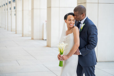 Groom kissing his bride's temple, standing behind her. as she smiles with her eyes closed holding a bouquet of calla lilies. Photograph is taken outdoors in an urban area with large columns.