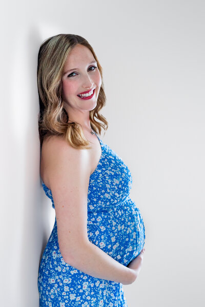A pregnant woman leans against the wall and smiles at the  camera while posing for her maternity pictures  in the studio