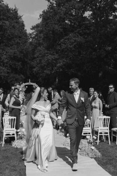 Een bruidspaar loopt hand in hand weg van het altaar waar zij zojuist getrouwd zijn. Hun gasten kijken hen liefdevol na. Deze foto is in zwart wit en vastgelegd door Sharon Kastelijns Photography