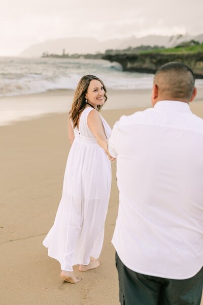 A woman looks back as she's holding the hands to her husband on the beach of Oahu.