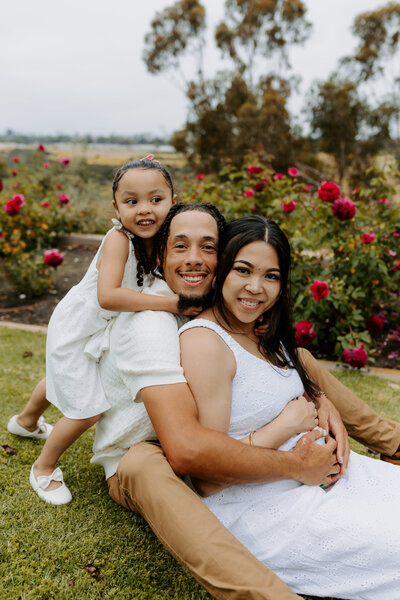 A smiling young family sits together in a rose garden, with their daughter climbing on dad’s back, all dressed in soft neutral tones.