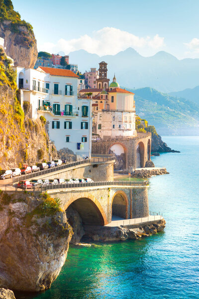 A picturesque coastal village with colorful buildings perched on a rocky cliff, overlooking a clear blue sea. Arched bridges connect the structures. Mountains are visible in the background under a sunny sky.