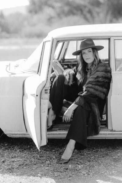 Wedding planner, Jessica Kuipers, wears fur stole and hat while sitting in drivers seat of vintage car