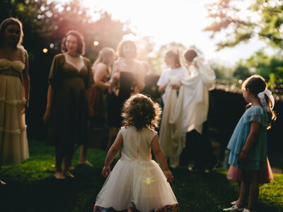 A whimsical outdoor wedding scene with a small child in a white dress among guests at sunset.