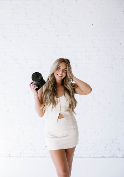 A woman photographer in a white outfit holds a camera, smiling against a white brick wall. She exudes a cheerful and confident vibe.